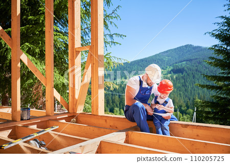 Dad with toddler son constructing wooden frame house. Male builder showing his son the construction plan, wearing helmets and blue overalls on sunny day. Carpentry and family concept. Dad with toddler son constructing wooden frame house. Male builder showing his son the construction plan, wearing helmets and blue overalls on sunny day. Carpentry and family concept. 110205725