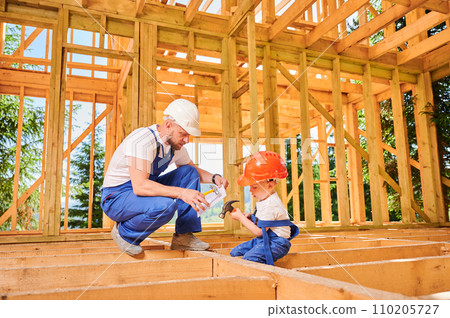 Father with toddler son building wooden frame house. Male builder teaching his son the technique of pounding nails with hammer on construction site, wearing helmets and blue overalls. 110205727