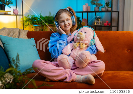 Happy child girl in wireless headphones relaxing sitting on sofa at home listening favorite music 110205836