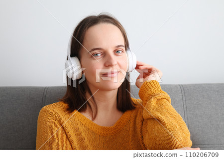Beautiful attractive cute relaxed woman listening to music with headphones sitting on sofa at home enjoying favorite playlist looking at camera with smile 110206709