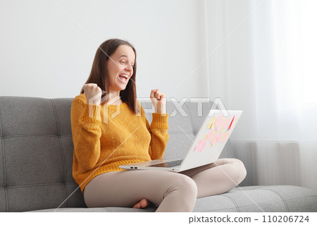 Website surfing and technology. Digital business and online operations. Overjoyed Caucasian woman using laptop sitting on sofa at home clenched fists rejoicing celebrating success Website surfing and technology. Digital business and online operations. Overjoyed Caucasian woman using laptop sitting on sofa at home clenched fists rejoicing celebrating success 110206724