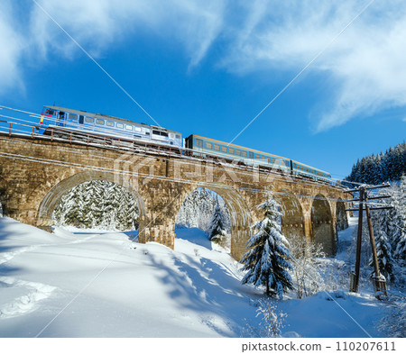 Stone viaduct (arch bridge) on railway through mountain snowy fir forest. Snow drifts  on wayside and hoarfrost on trees and electric line wires. 110207611