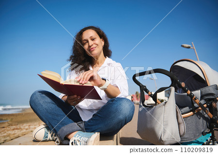 Portrait of authentic mother reading book outdoor, spending nice time on the beach with her baby sleeping in stroller 110209819