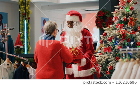 Customers in xmas adorn clothing store giving raffle ticket to worker dressed as Santa Claus holding gift acting as collecting box. Employee organizing winter holiday season promotional event 110210690