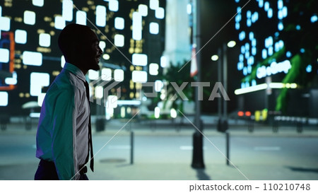 African american person on night walk downtown under streetlights, admiring modern office buildings from illuminated sidewalk. Confident man enjoying nighttime promenade around town. 110210748