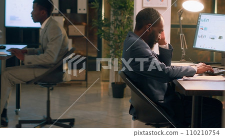 Overworked african american employee asleep in desk chair during nightshift, being woken up by fresh coworker. Joyful colleague playing jokes on sleepy businessman, scaring him 110210754