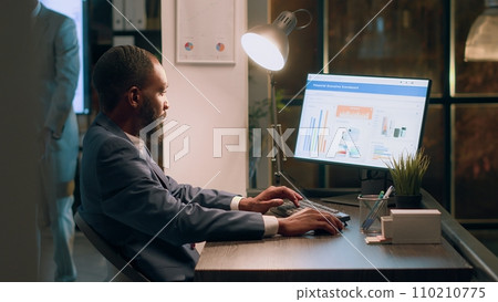 Experienced computer operator sitting at desk during nightshift, working on company accounting payroll reports and tables in modern office while coworker in background comes back from break Experienced computer operator sitting at desk during nightshift, working on company accounting payroll reports and tables in modern office while coworker in background comes back from break 110210775