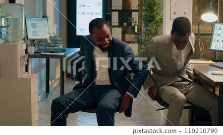 Colleagues laughing and having fun at work during nightshift, using desk chairs to race eachother in office hallways. Relaxed businessmen left unsupervised overnight playing together in workspace 110210790