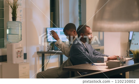 African american employee having fun at work during nightshift, bumping into coworker using desk chair. Friends racing eachother to avoid boredom during overnight stay in office 110210802