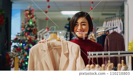 Portrait of happy woman browsing through elegant blazers on racks in Christmas adorn clothing store during winter holiday season. Smiling client checking fashion boutique items Portrait of happy woman browsing through elegant blazers on racks in Christmas adorn clothing store during winter holiday season. Smiling client checking fashion boutique items 110210803