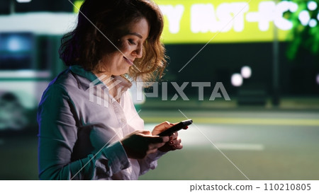 Smiling woman texting messages and walking around city center, reading texts and enjoying night time promenade. Person relaxing on evening stroll near skyscrapers under streetlights. Handheld shot. Smiling woman texting messages and walking around city center, reading texts and enjoying night time promenade. Person relaxing on evening stroll near skyscrapers under streetlights. Handheld shot. 110210805