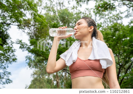 Young Asian woman in fitness clothes stops to drink water from her plastic water bottle during her morning exercise run at running track of a local park Young Asian woman in fitness clothes stops to drink water from her plastic water bottle during her morning exercise run at running track of a local park 110212861
