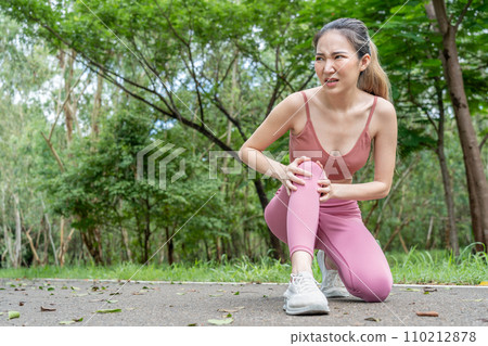 Young atractive Asian woman in fitness clothes with knee pain putting her hands over her troubled right knee while sitting down at running track of a local park 110212878