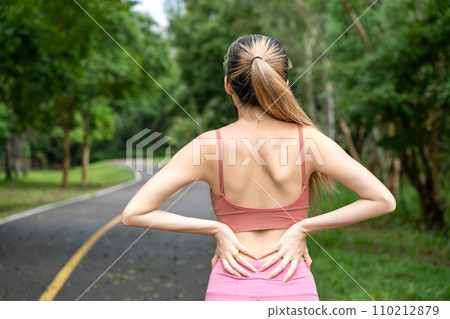 Back view of a long hair woman with back pain putting her hands over her trouble lower back while standing at running track of a local park 110212879