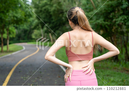 Back view of a long hair woman with back pain putting her hands over her trouble lower back while standing at running track of a local park 110212881