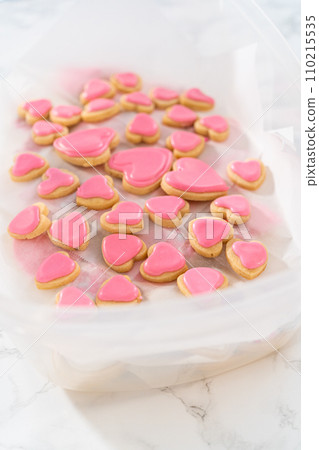 Storing heart-shaped sugar cookies with pink and white royal icing in a large plastic container. Storing heart-shaped sugar cookies with pink and white royal icing in a large plastic container. 110215535