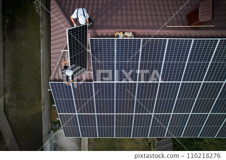 Men technicians mounting photovoltaic solar moduls on roof of house. Engineers in helmet installing solar panel system outdoors. Concept of alternative and renewable energy. Aerial view. Men technicians mounting photovoltaic solar moduls on roof of house. Engineers in helmet installing solar panel system outdoors. Concept of alternative and renewable energy. Aerial view. 110218276