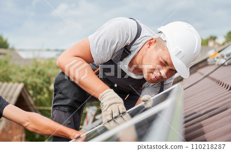 Men mounters mounting photovoltaic solar moduls on roof of house. Close up electrician in helmet looking on evenness of solar panel during installing. Concept of alternative and renewable energy. 110218287