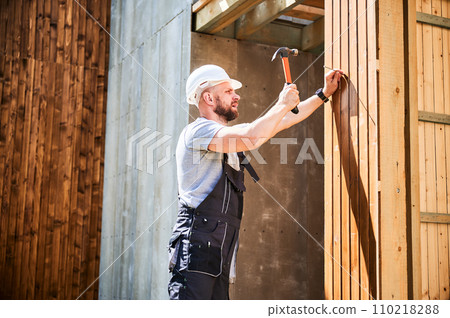 Carpenter constructing wooden frame, two-story house near forest. Bearded man hammering nails with hammer while wearing protective helmet and work coveralls. Concept of modern ecological construction. Carpenter constructing wooden frame, two-story house near forest. Bearded man hammering nails with hammer while wearing protective helmet and work coveralls. Concept of modern ecological construction. 110218288