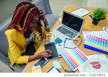 woman fashion designer with afro pigtails dreadlocks working on her atelier or print publishing house and graphic designer draws a sketch on a tablet. cosmetic distributor working in the office woman fashion designer with afro pigtails dreadlocks working on her atelier or print publishing house and graphic designer draws a sketch on a tablet. cosmetic distributor working in the office 110219013