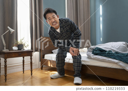 A man in his 50s in pajamas holding his knees next to the bed 110219333