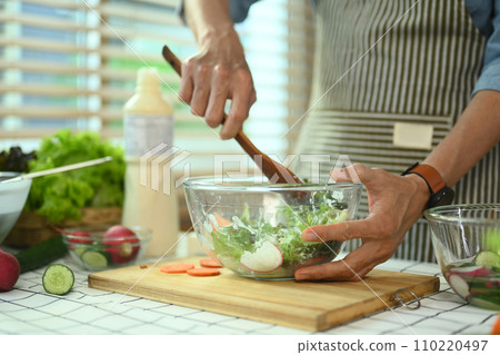Cropped shot of middle age man in apron mixing fresh organic vegetables in glass bowl. Healthy eating concept Cropped shot of middle age man in apron mixing fresh organic vegetables in glass bowl. Healthy eating concept 110220497