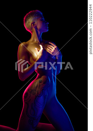 Attractive young woman with short blonde hair, slim fit body posing naked isolated over black background in neon light Attractive young woman with short blonde hair, slim fit body posing naked isolated over black background in neon light 110222944