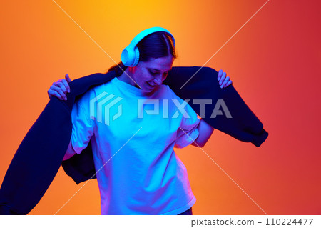 Young attractive woman with freckles dressed stylish outfit listening music in head phones against gradient orange-yellow background in neon light. 110224477