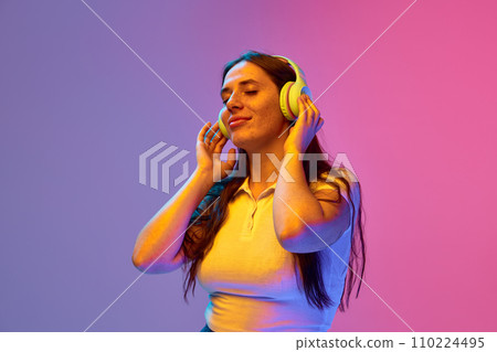 Positive young woman, student enjoying, dancing while listening music in headphones against gradient studio background in neon light, filter. Positive young woman, student enjoying, dancing while listening music in headphones against gradient studio background in neon light, filter. 110224495