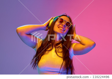 Positive, overjoyed girl, student singing and dancing while listening music in headphones against gradient studio background in neon light, filter. Positive, overjoyed girl, student singing and dancing while listening music in headphones against gradient studio background in neon light, filter. 110224496