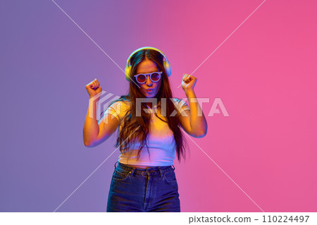 Positive, overjoyed girl, student dancing while listening music in headphones against gradient studio background in neon light, filter. Positive, overjoyed girl, student dancing while listening music in headphones against gradient studio background in neon light, filter. 110224497