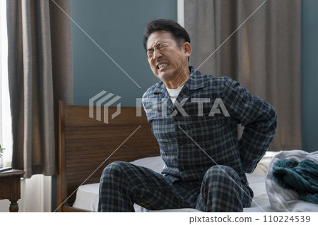 Image of a man in his 50s sitting on the bed with his hands on his hips, back pain, stiff lower back 110224539