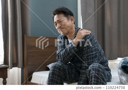 Image of a man in his mid-50s sitting on the bed and holding his shoulders, stiff shoulders, frozen shoulders 110224738