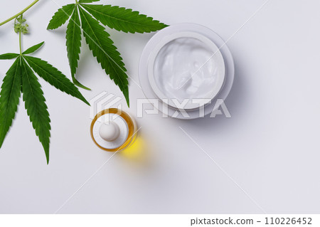 Container of hemp cream and CBD oil top view on white background. Cannabis body care cosmetics with marijuana leaf. 110226452