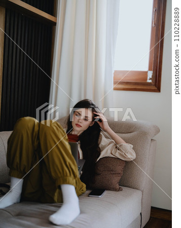 A woman lies on a couch with a laptop and stares pensively into the screen training a freelancer at work, a real lifestyle without retouching. 110228889