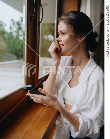 Beautiful woman with phone in hand standing at home window, freelancer on vacation, home convenient lifestyle with online work, cyber security concept, smart home. 110228900