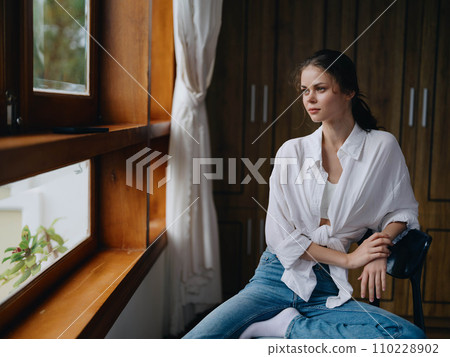 Woman sitting at home by a wooden window with a smile on a chair in homemade comfortable clothes and looking at the landscape, spring mood, women's day, rest on the weekend. 110228902