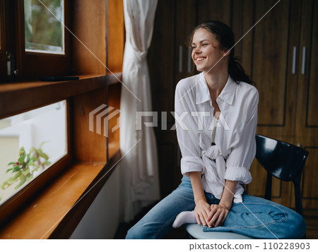Woman sitting at home by a wooden window with a smile on a chair in homemade comfortable clothes and looking at the landscape, spring mood, women's day, rest on the weekend. 110228903