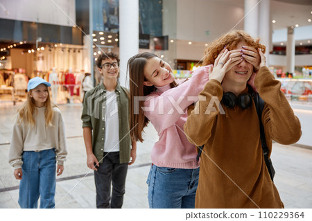 Cute overjoyed girl covering eyes of boy while friends walking in shopping mall Cute overjoyed girl covering eyes of boy while friends walking in shopping mall 110229364