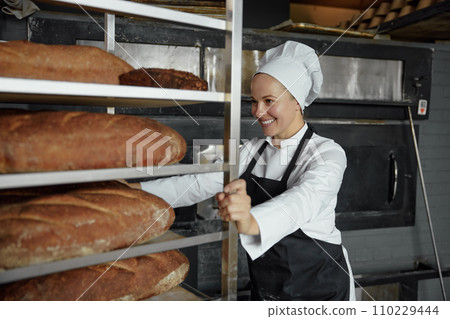Woman baker smiling happily pushing rack with fresh baked bread 110229444