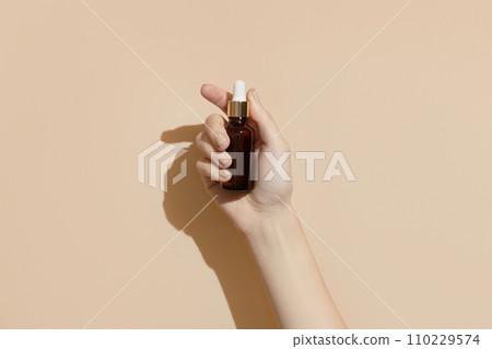 A womans hand holding a brown bottle of serum on a beige background. Body care  110229574