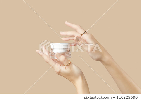 A womans hands holding a white cosmetic jar on a beige background. Beauty treatment concept A womans hands holding a white cosmetic jar on a beige background. Beauty treatment concept 110229590