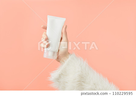 A womans hand in a sweater holds a tube of cream on a peach background. Self care beauty treatment 110229595