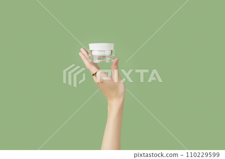 A womans hand holding a white cosmetic jar on a green background 110229599