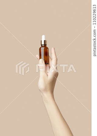 A womans hand holding a brown bottle of serum on a beige background. Body care A womans hand holding a brown bottle of serum on a beige background. Body care 110229613