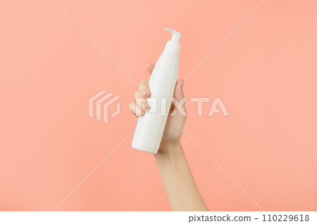 A womans hand holding a white cosmetic bottle on a peach background. Beauty treatment concept A womans hand holding a white cosmetic bottle on a peach background. Beauty treatment concept 110229618