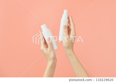 A womans hands holding a white cosmetic bottles on a peach background. Beauty treatment concept A womans hands holding a white cosmetic bottles on a peach background. Beauty treatment concept 110229620
