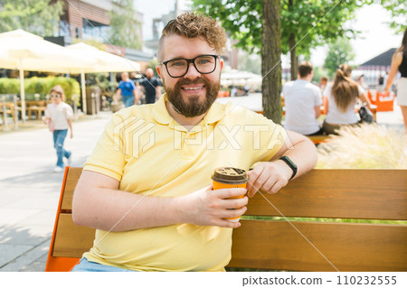 Millennial smiling student man in yellow polo t-shirt eyeglasses sitting on bench drink coffee have breakfast rest relax in sunshine spring green city park outdoors on nature. Urban leisure concept. 110232555