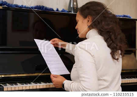 Rear view female pianist sits at piano and reads music notes, ready to perform classical musical melody on grand piano Rear view female pianist sits at piano and reads music notes, ready to perform classical musical melody on grand piano 110233515