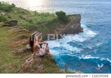 A beautiful woman in a pink dress sits on a cliff above the ocean on the island of Nusa Penida. Devil's Billabong an incredibly wonderful lagoon with splashes from the waves. A beautiful woman in a pink dress sits on a cliff above the ocean on the island of Nusa Penida. Devil's Billabong an incredibly wonderful lagoon with splashes from the waves. 110234308